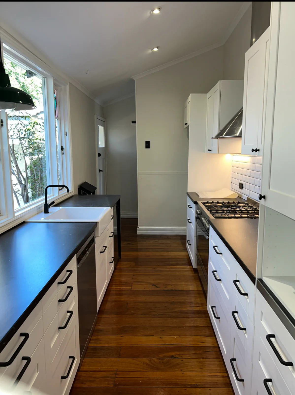 Clean galley kitchen with white shaker cabinetry, a dark benchtop, a farmhouse sink and polished timber floorboards