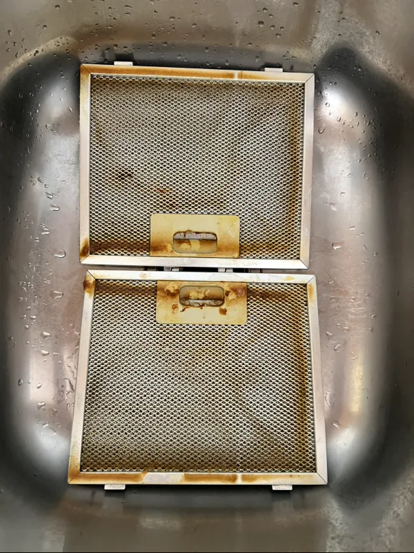 Two rangehood mesh filters coated in brown grease build up, lying in a stainless steel sink before cleaning